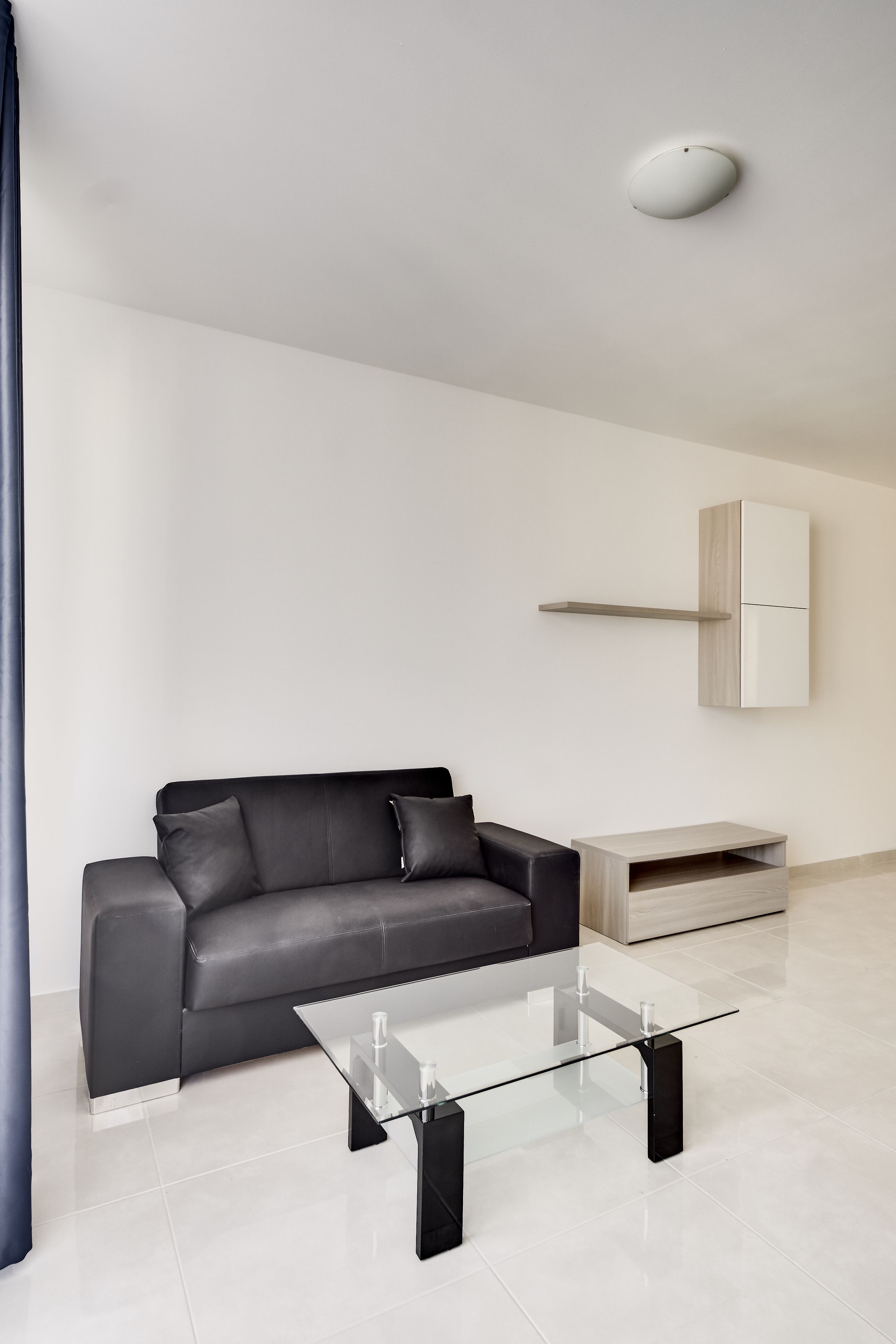 A modern living room with a black leather sofa, a glass coffee table, a wooden cabinet, and a white ceiling light.