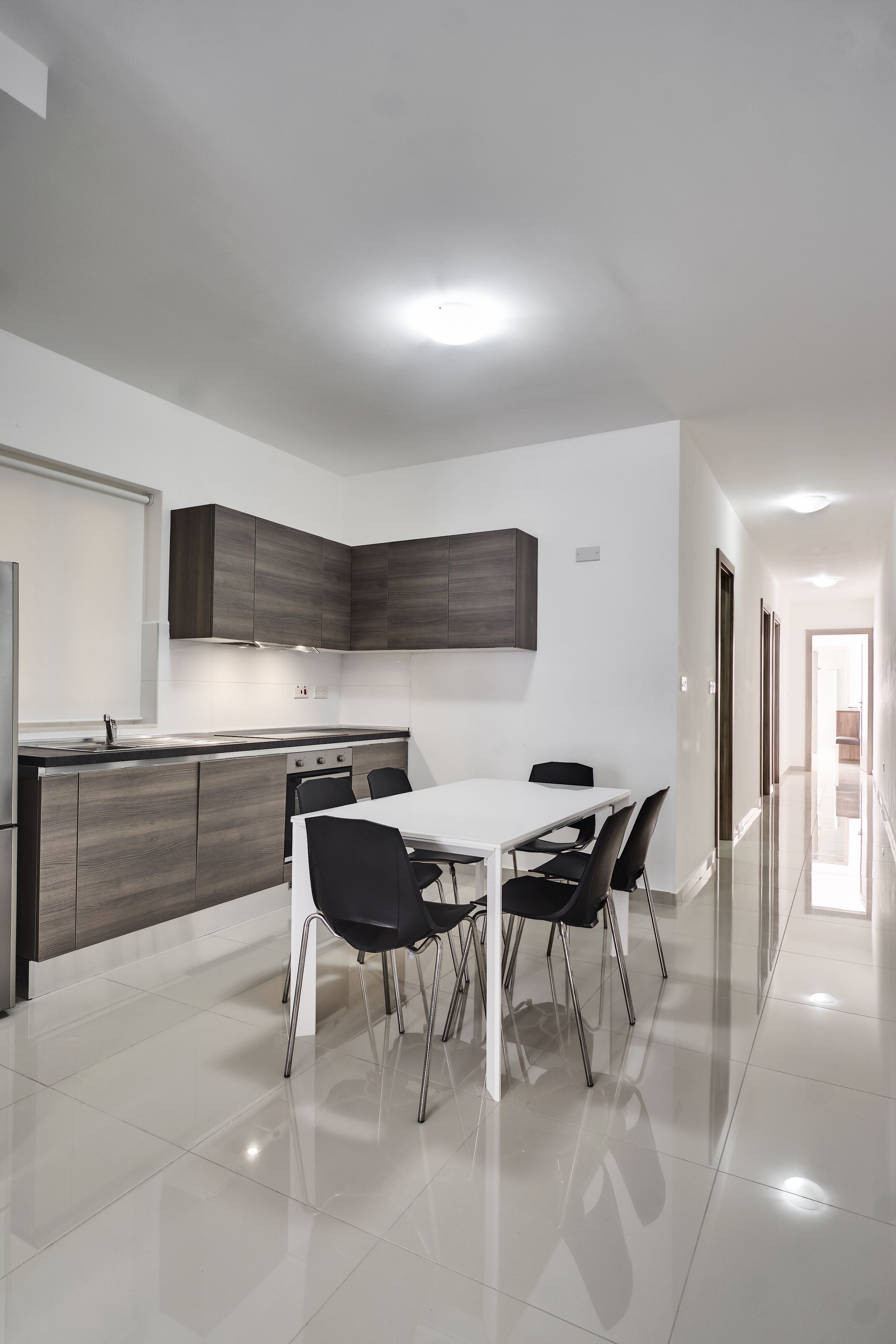A modern kitchen with a white dining table and black chairs, a stainless steel refrigerator, and dark wooden cabinets.