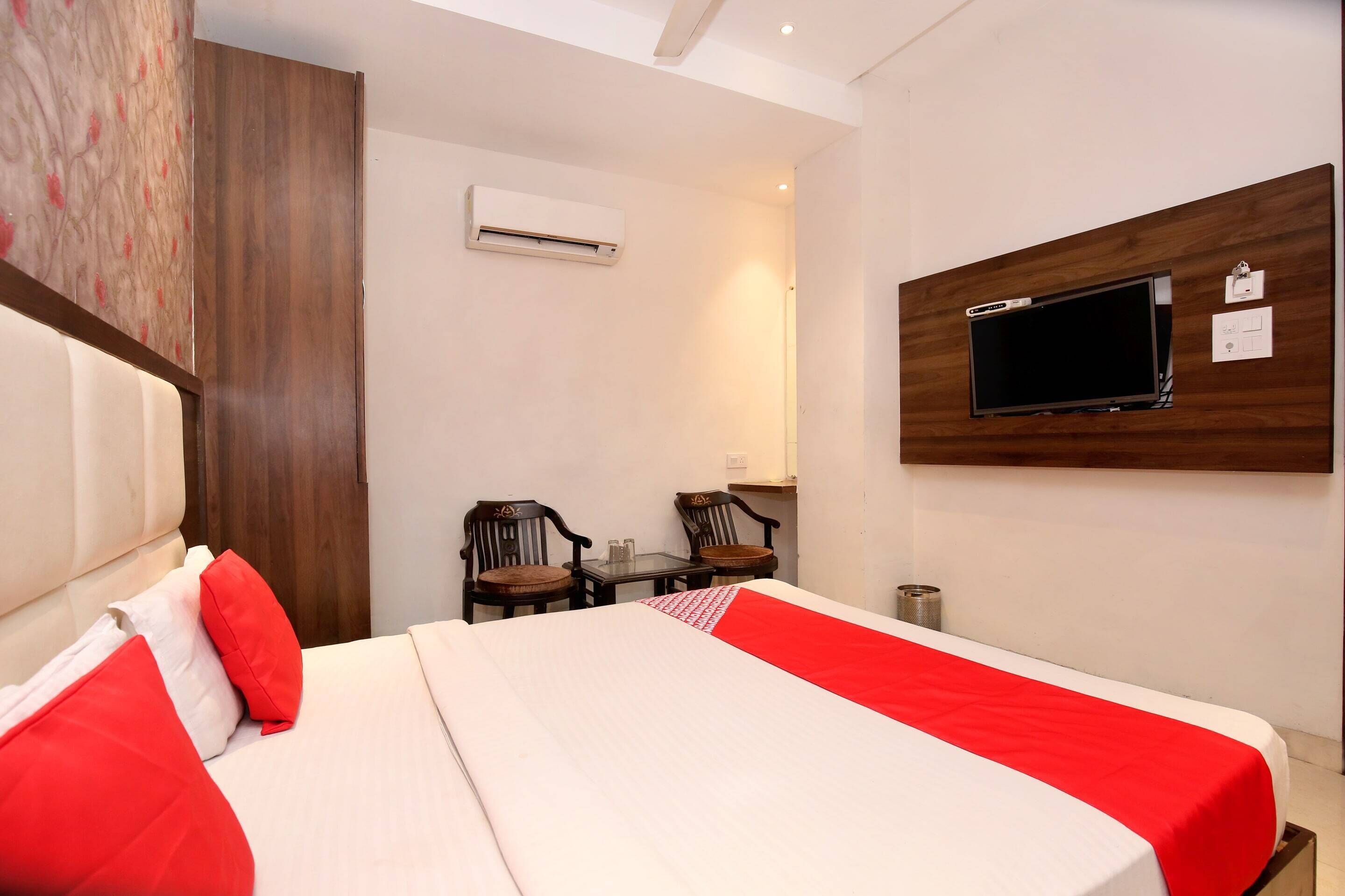 A hotel room with a bed, two chairs, a small table, an air conditioning unit, and a mounted TV.