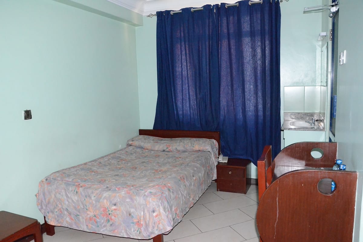 Quarto com cama, cadeira de madeira, mesa de cabeceira e banheiro visível por uma cortina.
