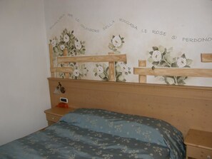 A bedroom with a wooden headboard, a bed with floral bedding, a nightstand, and a wall with floral wallpaper. - Villa Ilaria (Caderzone Terme)