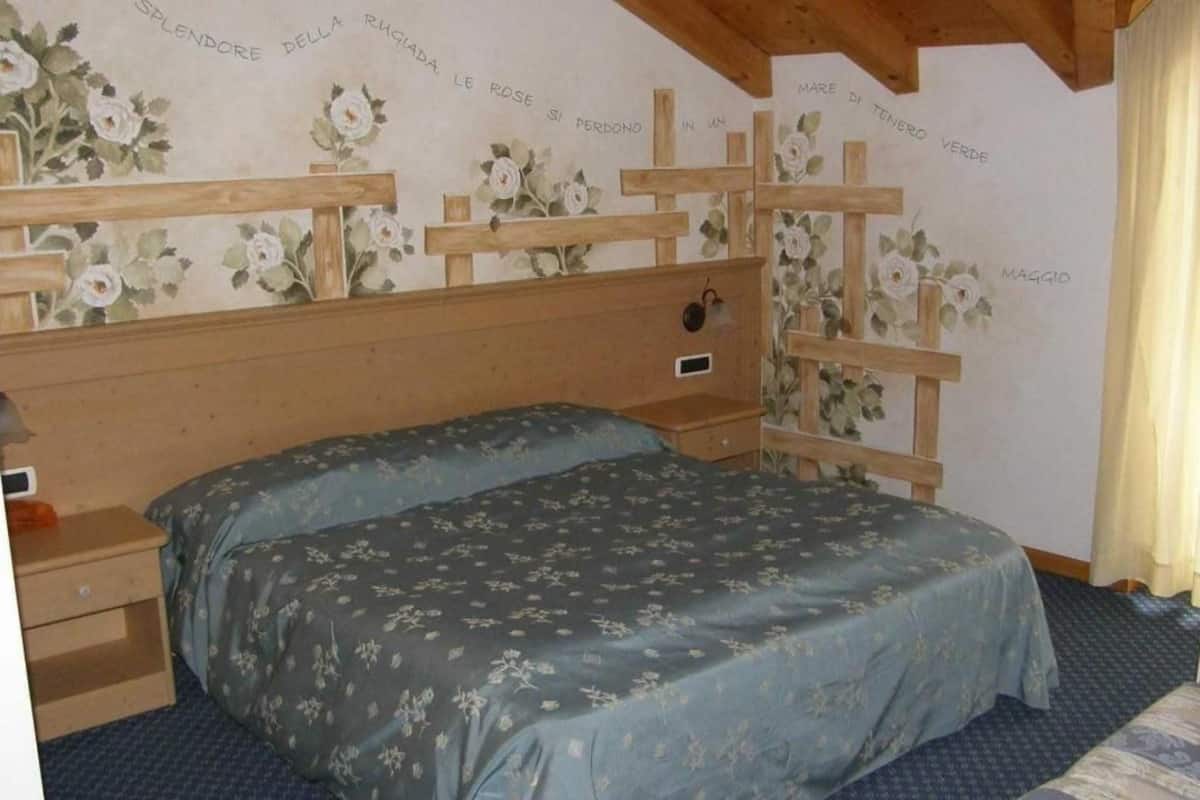 A bedroom with a wooden headboard, floral wallpaper, a bed with a floral bedspread, a nightstand, and a small wooden shelf.