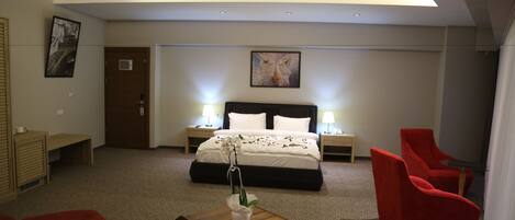 A hotel room with a bed, two bedside tables with lamps, a wooden coffee table with a vase, a red armchair, and a red sofa.