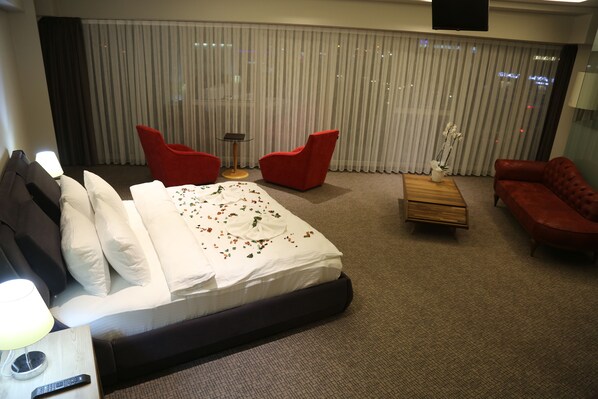 A hotel room with a bed, two red chairs, a red sofa, a small wooden table, and a large window with curtains.