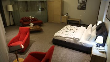 Una habitación de hotel con una cama, un sillón rojo, un sofá rojo, una mesita, una mesita de noche con una lámpara y un cuadro en la pared.