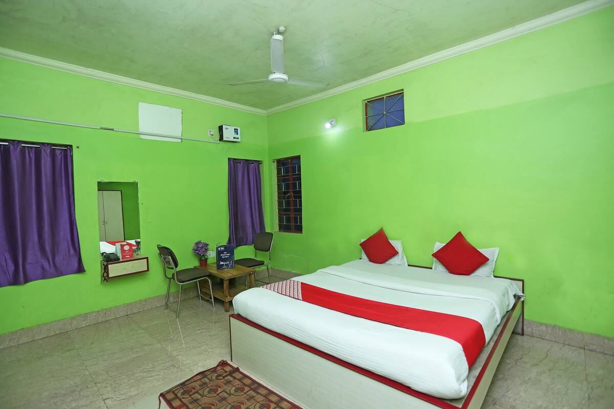 A room with green walls, a bed with red and white bedding, a ceiling fan, a desk with a chair, and a small window with bars.