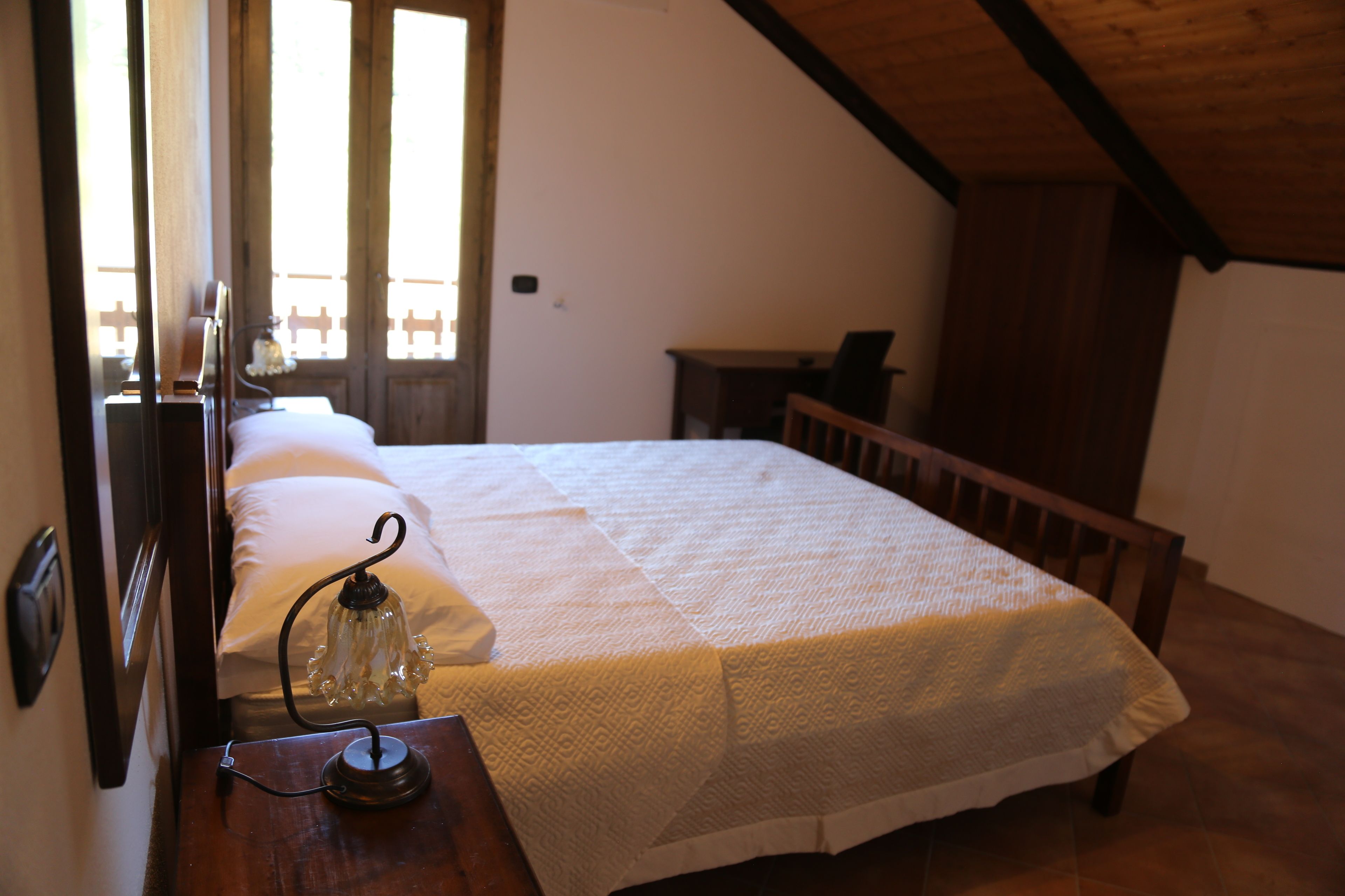 Un dormitorio con cama, una mesita de noche con lámpara, un banco de madera y una ventana con vistas a una valla.