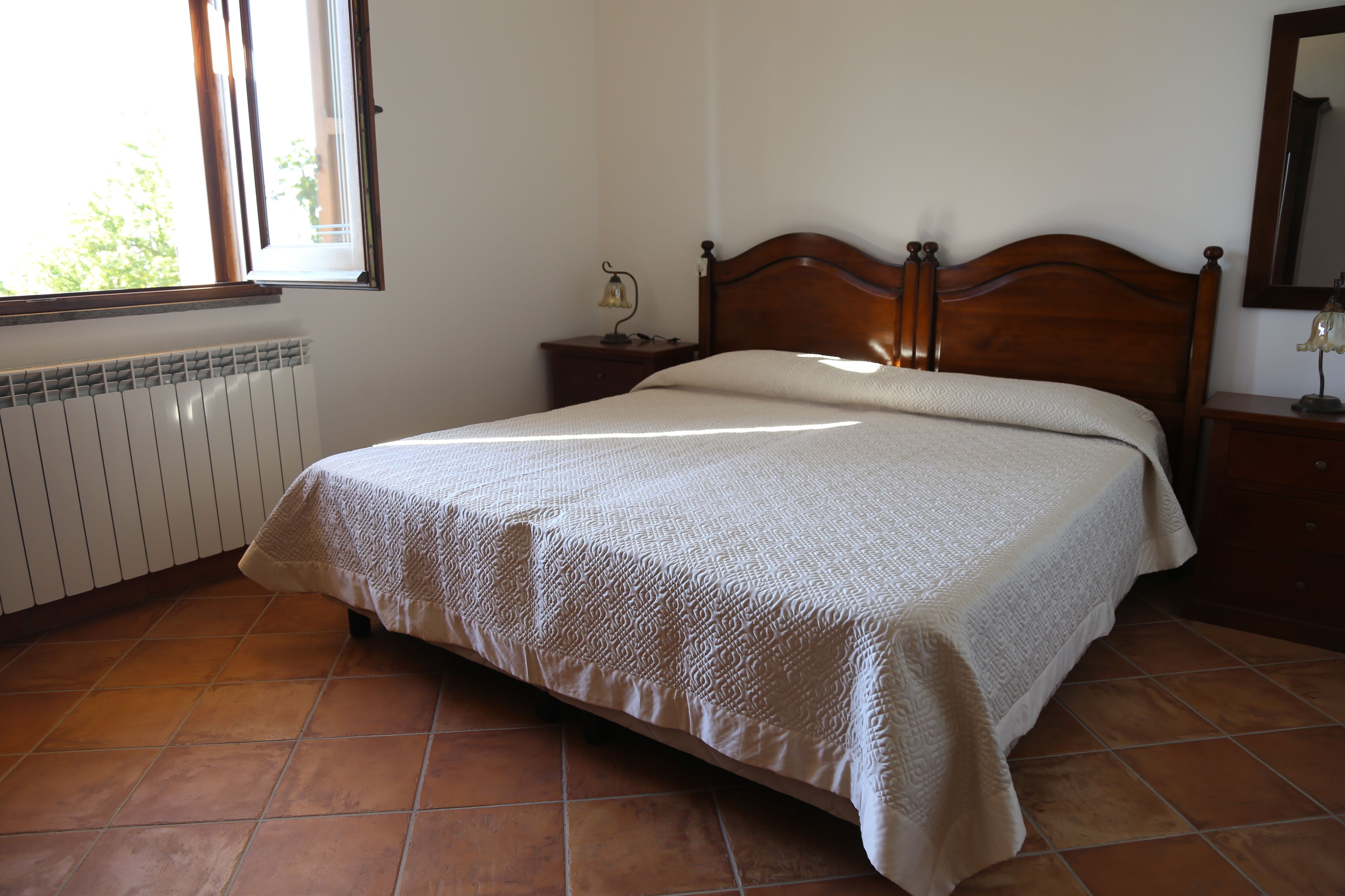 Quarto com cama de madeira, mesa de cabeceira com abajur, radiador e janela com persianas.