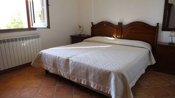 Quarto com cama de madeira, mesa de cabeceira com abajur, radiador e janela com persianas.