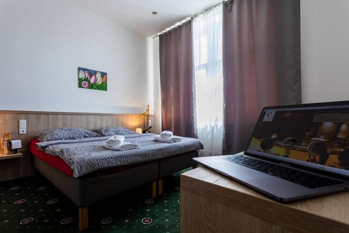 A hotel room with a bed, a laptop on a desk, a painting on the wall, and a window with curtains.