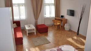 A compact living space with a red sofa, a small white table, a wooden desk with a laptop, a TV on the wall, and a decorative vase. - Penzion U Vlka (Ceske Budejovice)