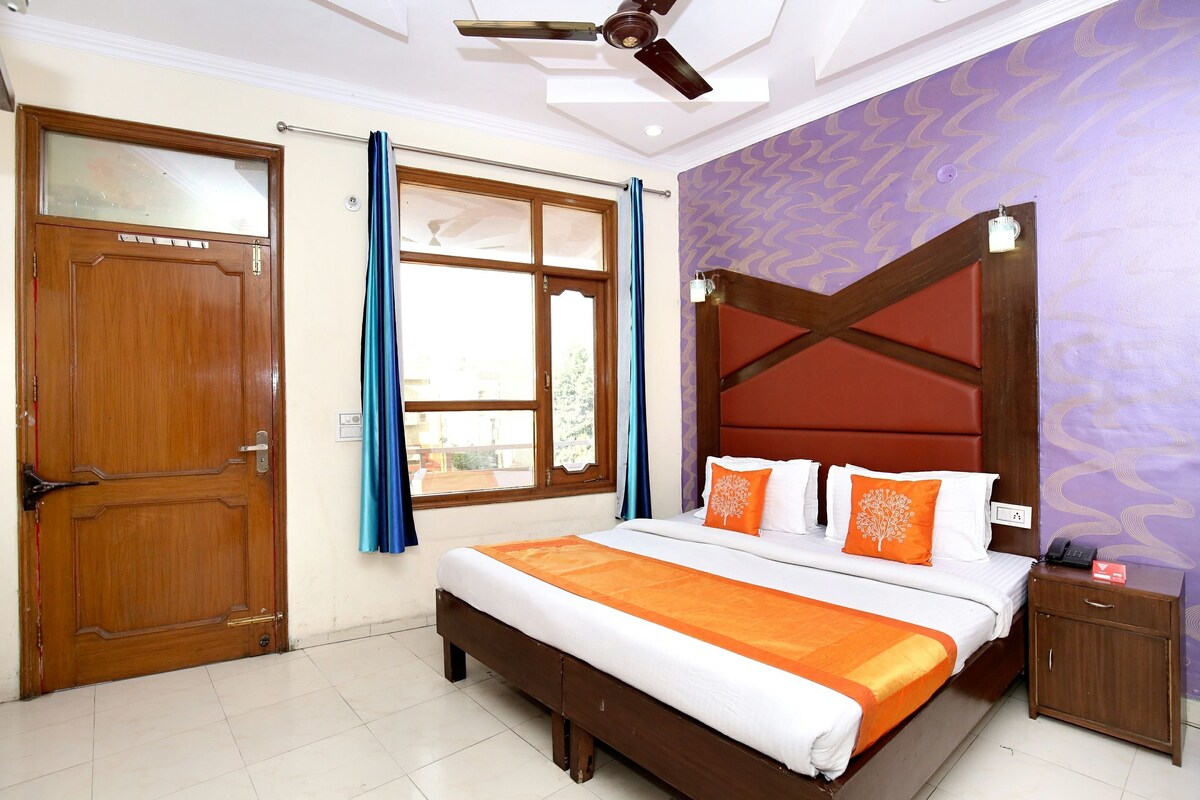 A hotel room with a wooden door, a large bed with an orange bedspread, a nightstand with a phone, and a ceiling fan.
