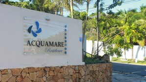 A sign for Acquamare Residence with a logo, a stone wall, and a paved area with trees and a fence in the background. - Pousada Acquamare (Búzios)