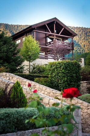 A wooden cabin with a stone foundation, surrounded by lush greenery and a mountainous backdrop. - Aelia Hospitality Parnassos (Amfikleia-Elateia)