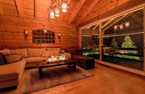 A cozy log cabin interior with a wooden ceiling, a large window with a view of a green landscape, a sofa, and a coffee table with candles. - Aelia Hospitality Parnassos (Amfikleia-Elateia)