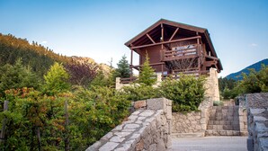 Eine Holzhütte mit einer Steintreppe, die zu einem Balkon führt, umgeben von üppiger Vegetation und Bergen.