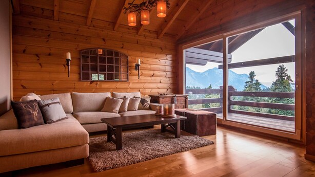 Ein gemütliches Blockhäuschen-Interieur mit Holzbalkendecke, einem großen Fenster mit Blick auf die Berge, einer Couch und einem Couchtisch.