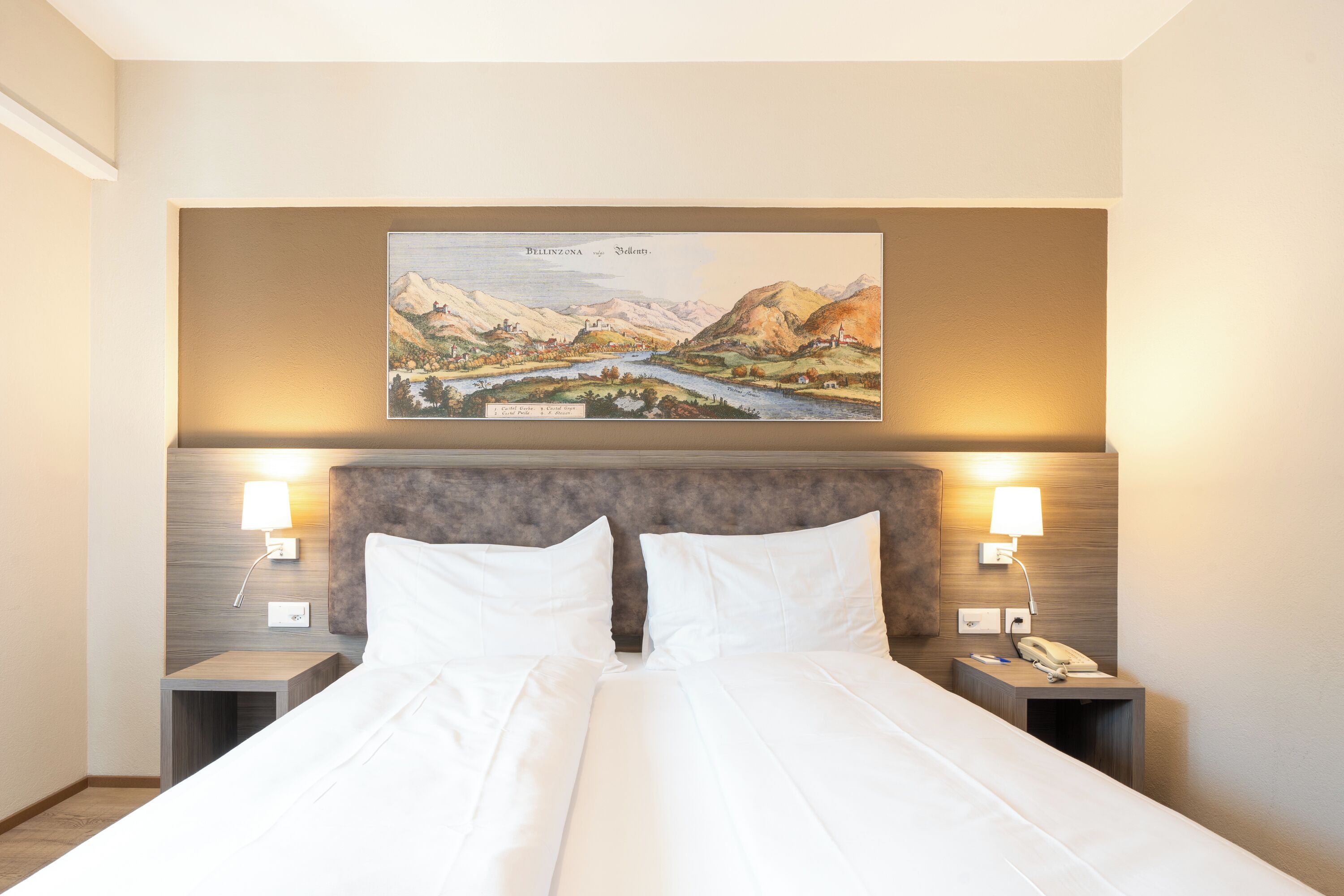 A hotel room with a large bed, two bedside tables, a headboard, and a wall-mounted painting of a landscape.