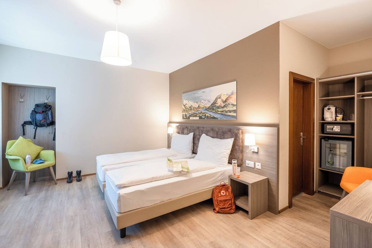 A hotel room with two beds, a green chair, a wooden wardrobe, and a painting on the wall.