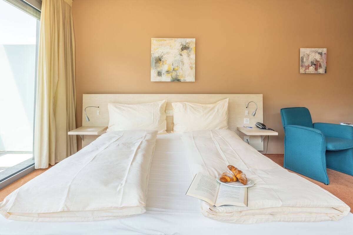A hotel room with a large bed, two bedside tables, a blue chair, and artwork on the walls.