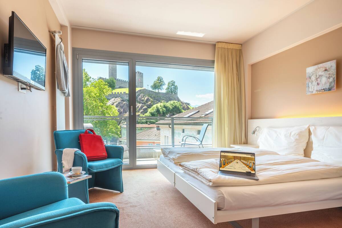 A hotel room with a bed, a TV, a laptop, a red bag, and a view of a castle through the window.