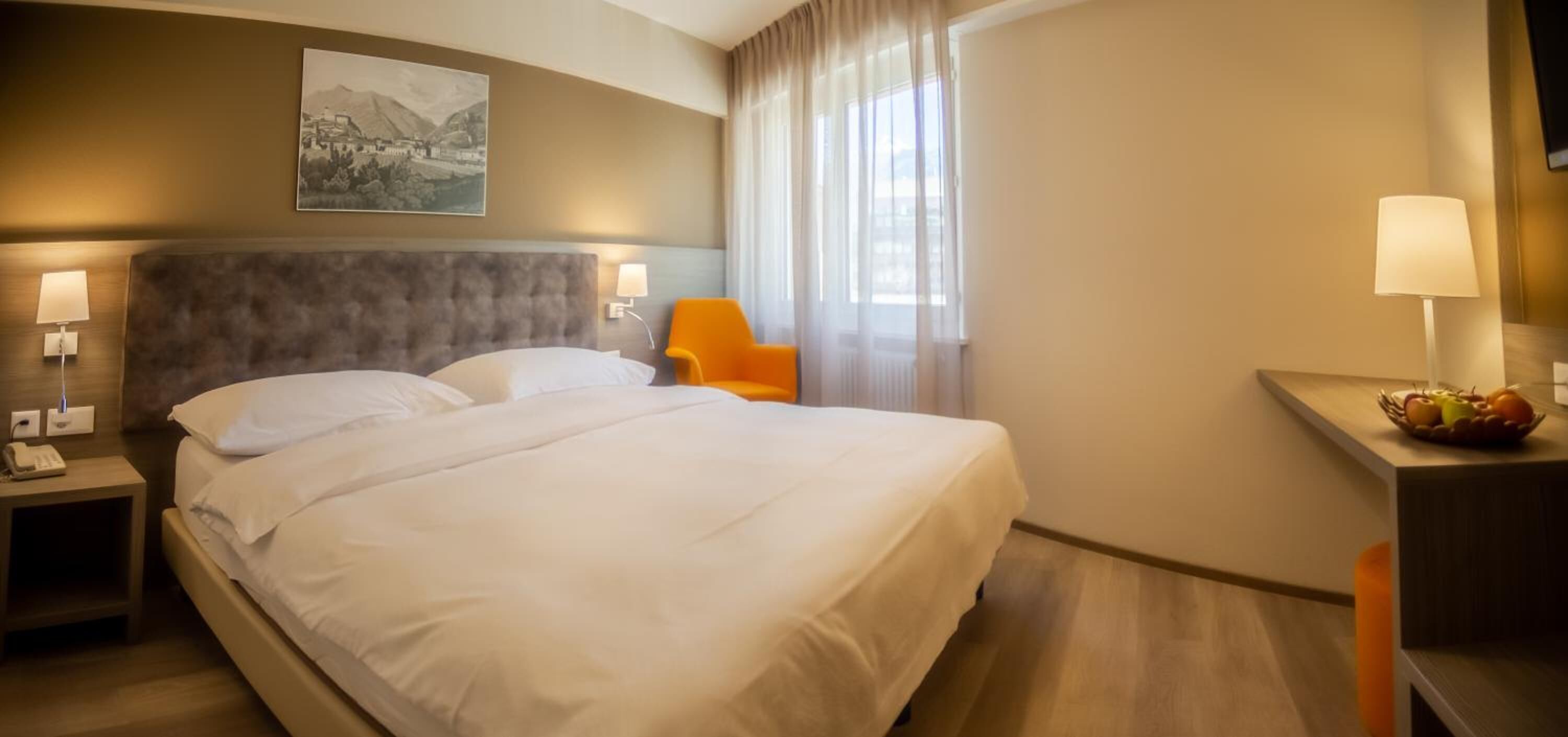 Et hotelværelse med en stor seng, to sengeborde med lamper, en stol, et skrivebord med lampe og en frugtkurv.