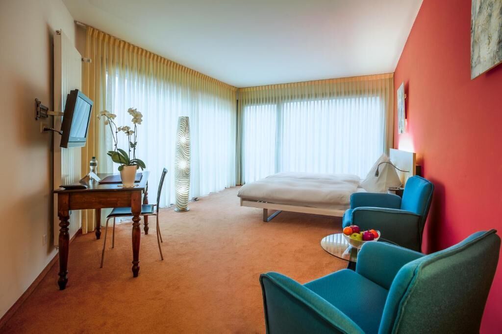 A hotel room with a bed, a desk with a computer, a chair, a TV, and a window with curtains.