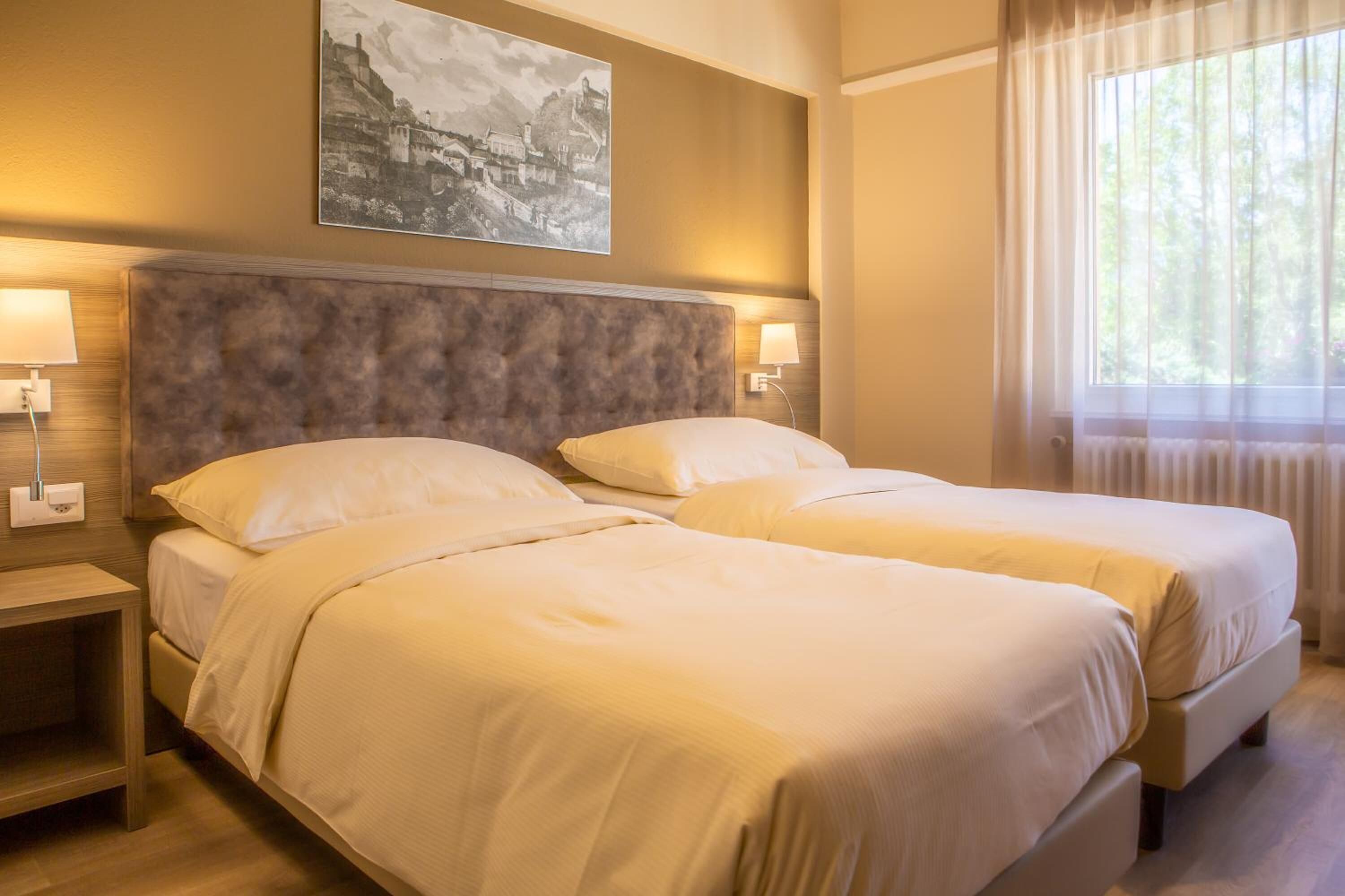 A hotel room with two beds, a headboard, bedside lamps, and a window with curtains.