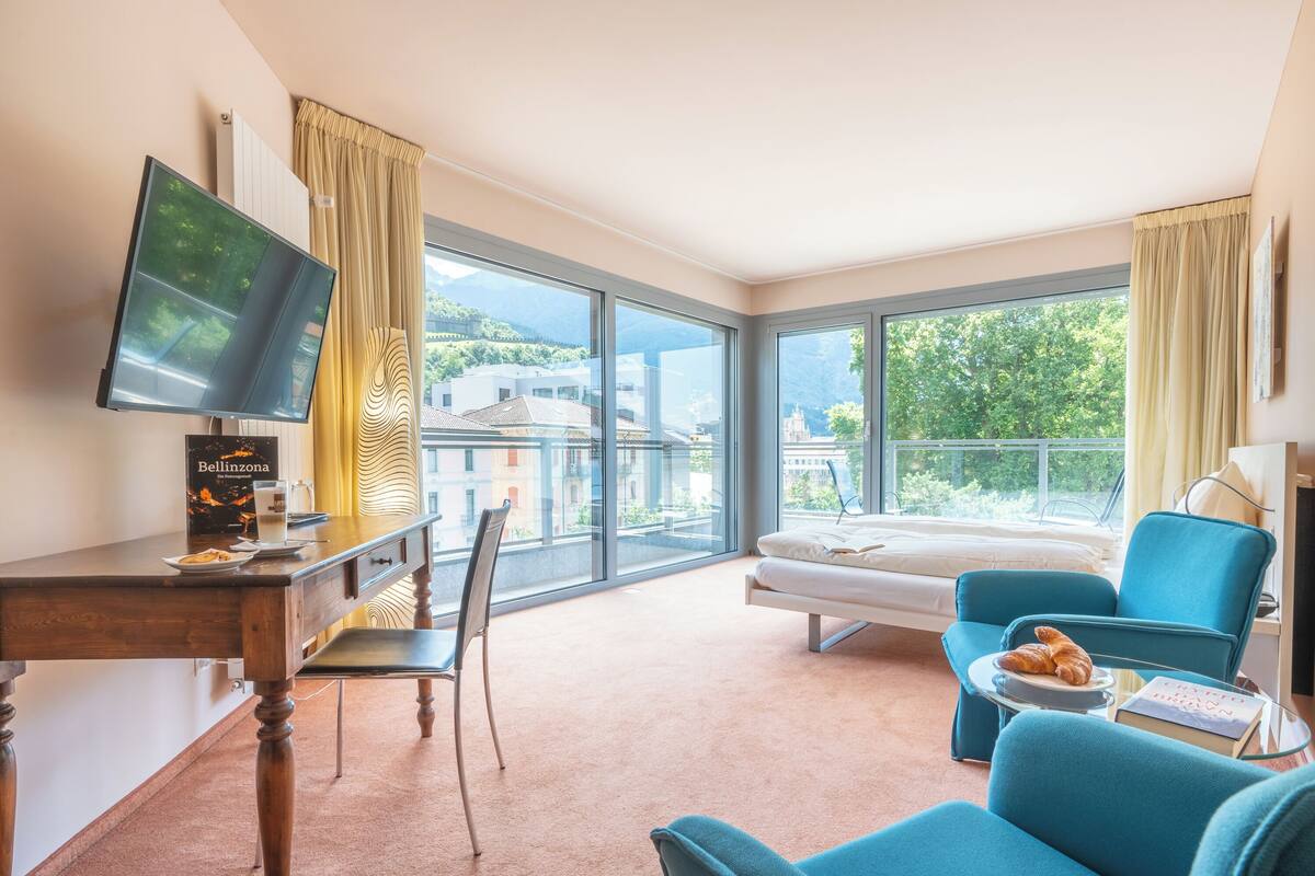 A hotel room with a large window, a bed, a desk with a computer, and a chair.