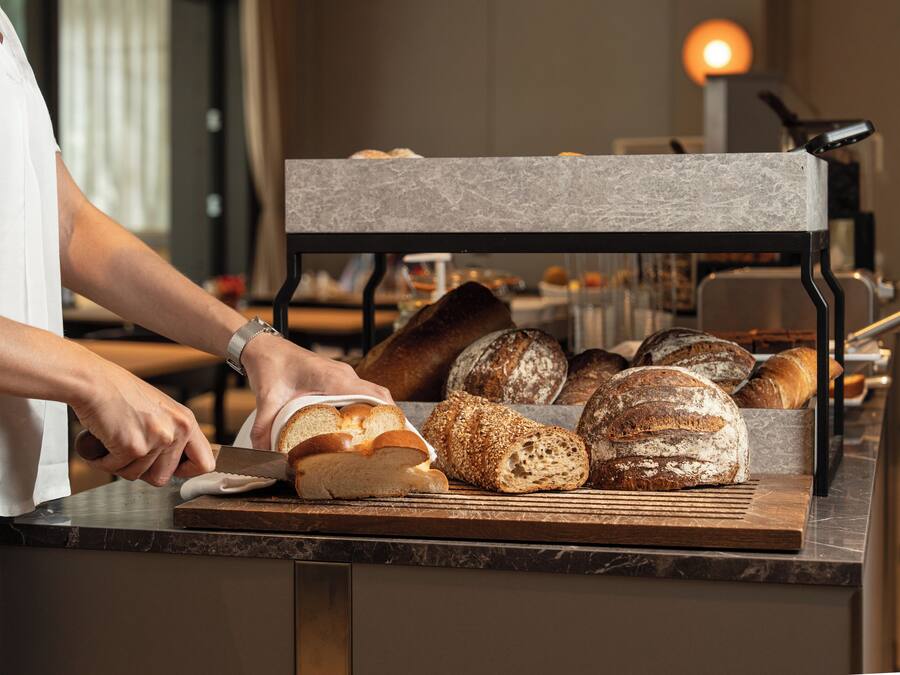 A person slicing bread on a marble countertop with various breads and pastries in the background.