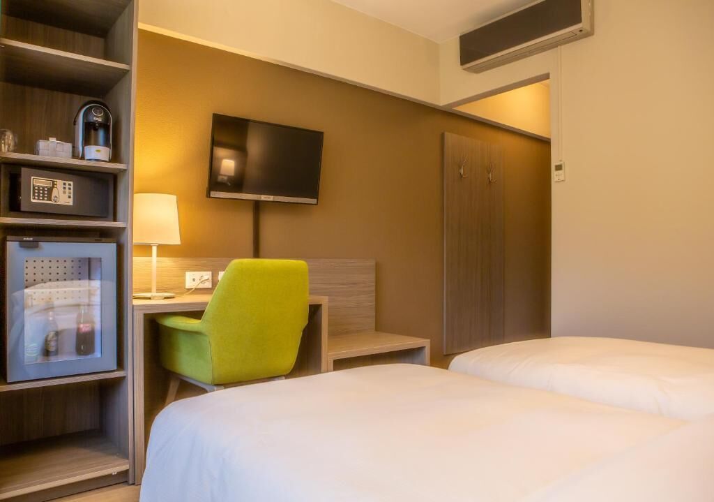 A hotel room with two beds, a desk with a lamp, a television, a minibar, and a green chair.