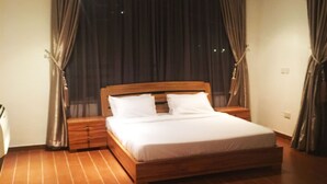 Uma cama grande e bem arrumada em um quarto espaçoso com piso de madeira e cortinas escuras.