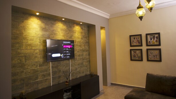 Uma sala de estar moderna com uma televisão de tela plana presa a uma parede texturizada, um console escuro, um sofá marrom e um lustre decorativo.