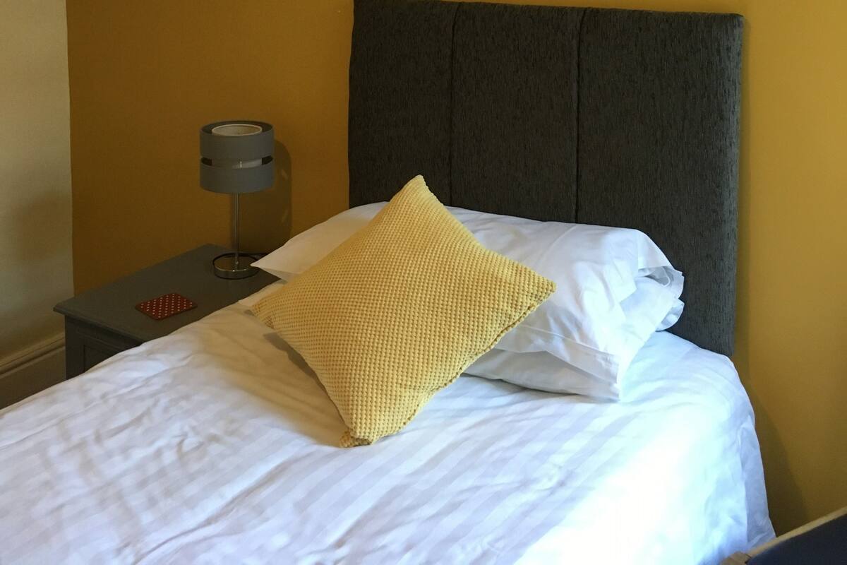 Una cama bien hecha con sábanas blancas y una única almohada amarilla, una mesita de noche gris con una lámpara y una pared lisa en un tono amarillo cálido.