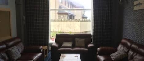 Um quarto com dois sofás de couro, uma mesa de centro e vista para um edifício através de uma janela com cortinas.