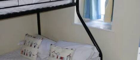 Ein Etagenbett mit weißer Bettwäsche und gemusterten Kissen, ein Fenster mit blauen Vorhängen und ein kleiner Nachttisch.