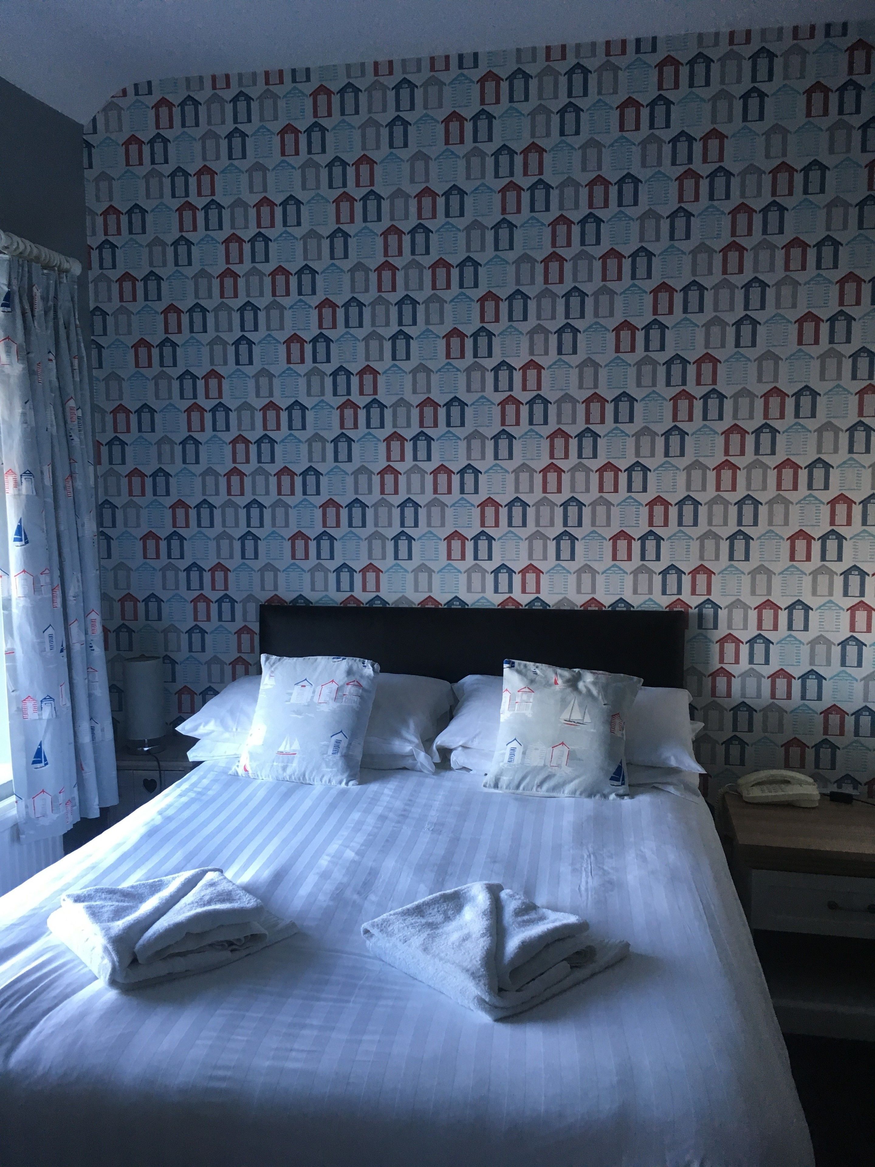A neatly made bed with white linens and pillows, a striped headboard, and a patterned wall with house designs.