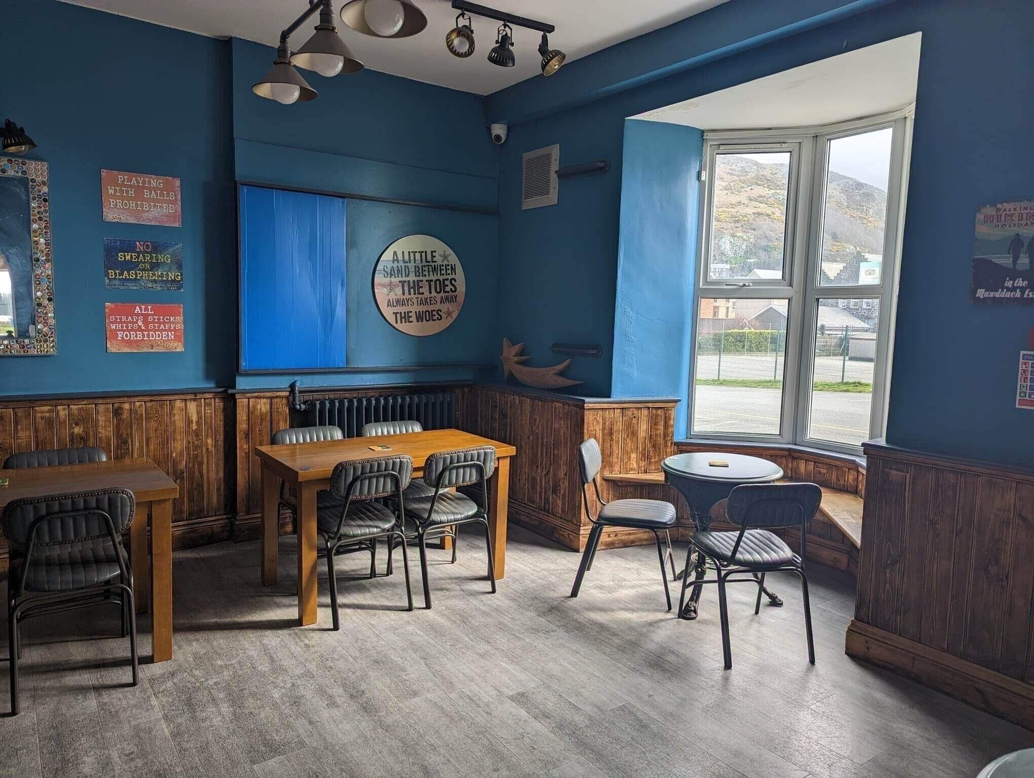 Interior de restaurante con pared azul, paneles de madera y un ventanal grande. Se observan mesas y sillas dispuestas para comer.