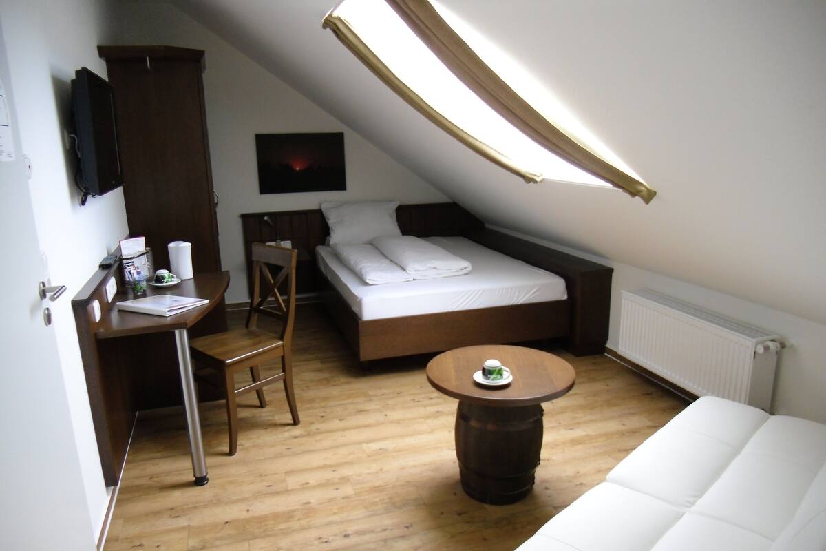 Une petite pièce, de taille modeste, comprenant un lit, un bureau et une chaise. On y trouve une fenêtre avec un rideau, un radiateur et une petite table sur laquelle est posée une tasse.