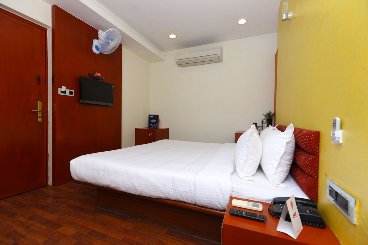 A hotel room with a wooden floor, a bed with white bedding, a bedside table with a remote control, a TV mounted on the wall, and an air conditioning unit.
