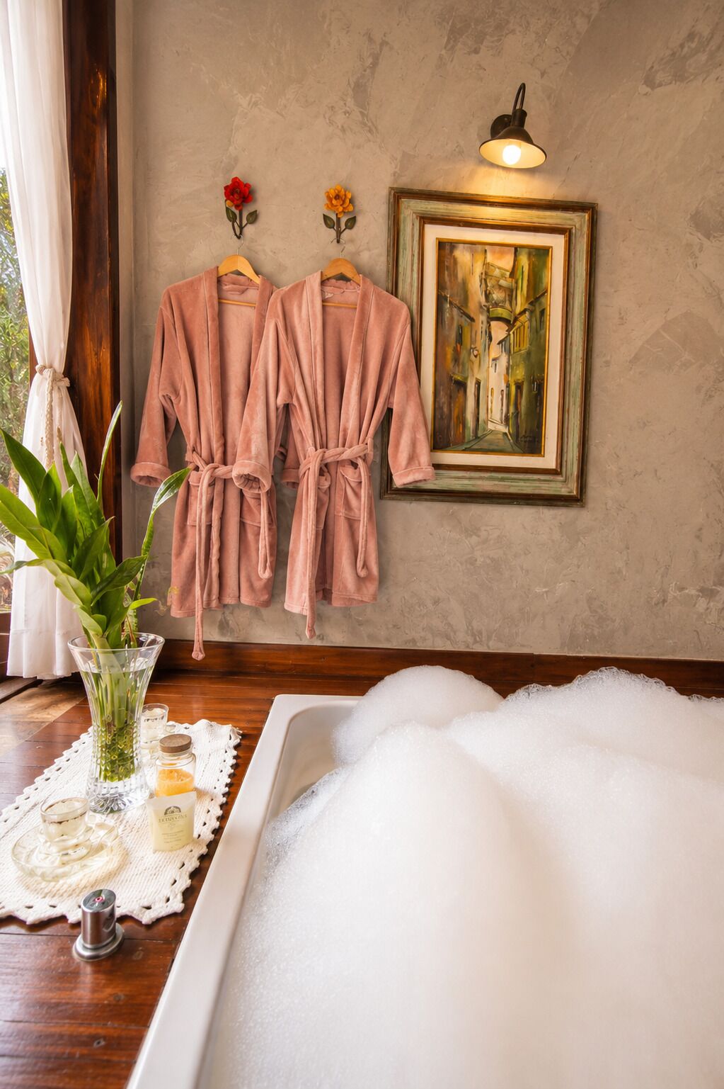 A spa room with a bathtub filled with bubbles, two pink robes hanging on hooks, a framed painting on the wall, and a potted plant on the windowsill.