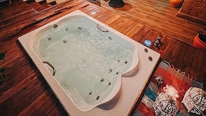 A spa room with a large hot tub, wooden flooring, a mirror, and a seating area with chairs and a small table. - Chalés Terraviva (Pirenópolis)