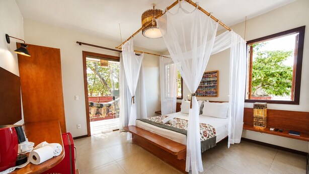 A room with a bed, a TV, a wooden bench, and a window with a view of greenery. - Chalés Terraviva (Pirenópolis)