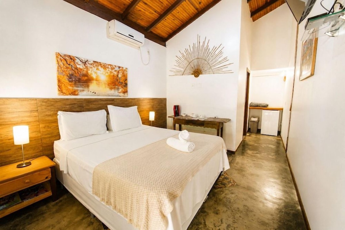 A hotel room with a large bed, a nightstand, a wall-mounted air conditioner, and a hallway leading to a bathroom.