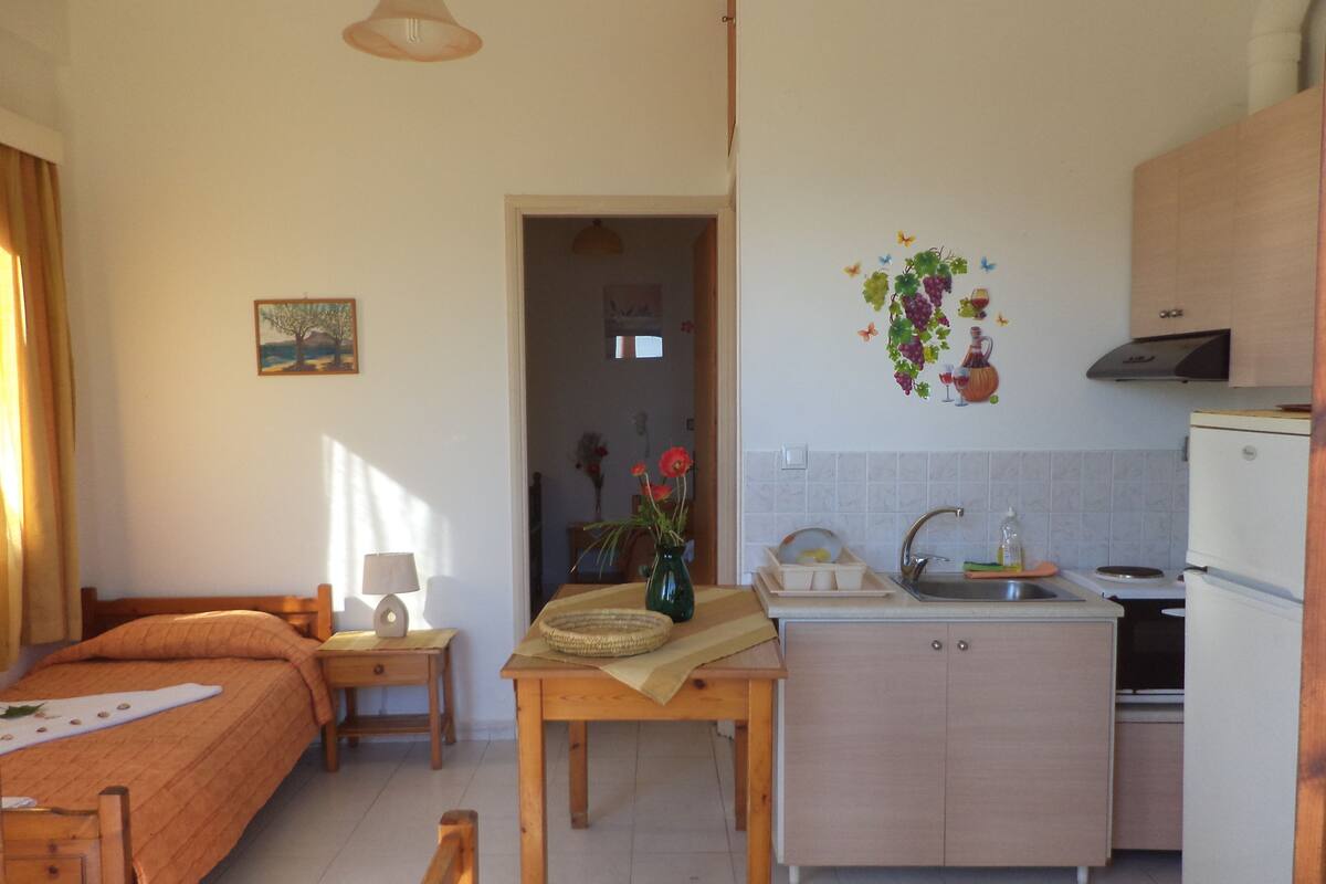 A compact room with a bed, a small table with a lamp, a kitchenette with a sink and a refrigerator, and a door leading to another room.
