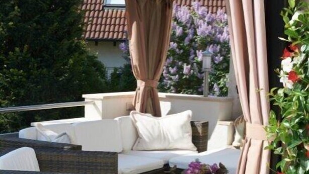 Un espace extérieur aménagé avec un canapé et des fauteuils en osier, un pot de fleurs et une pergola agrémentée de rideaux.