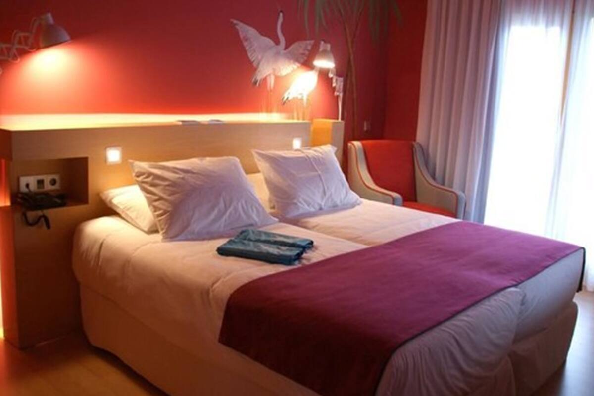 A hotel room with a large bed, a red wall, a white swan mural, and a red chair.