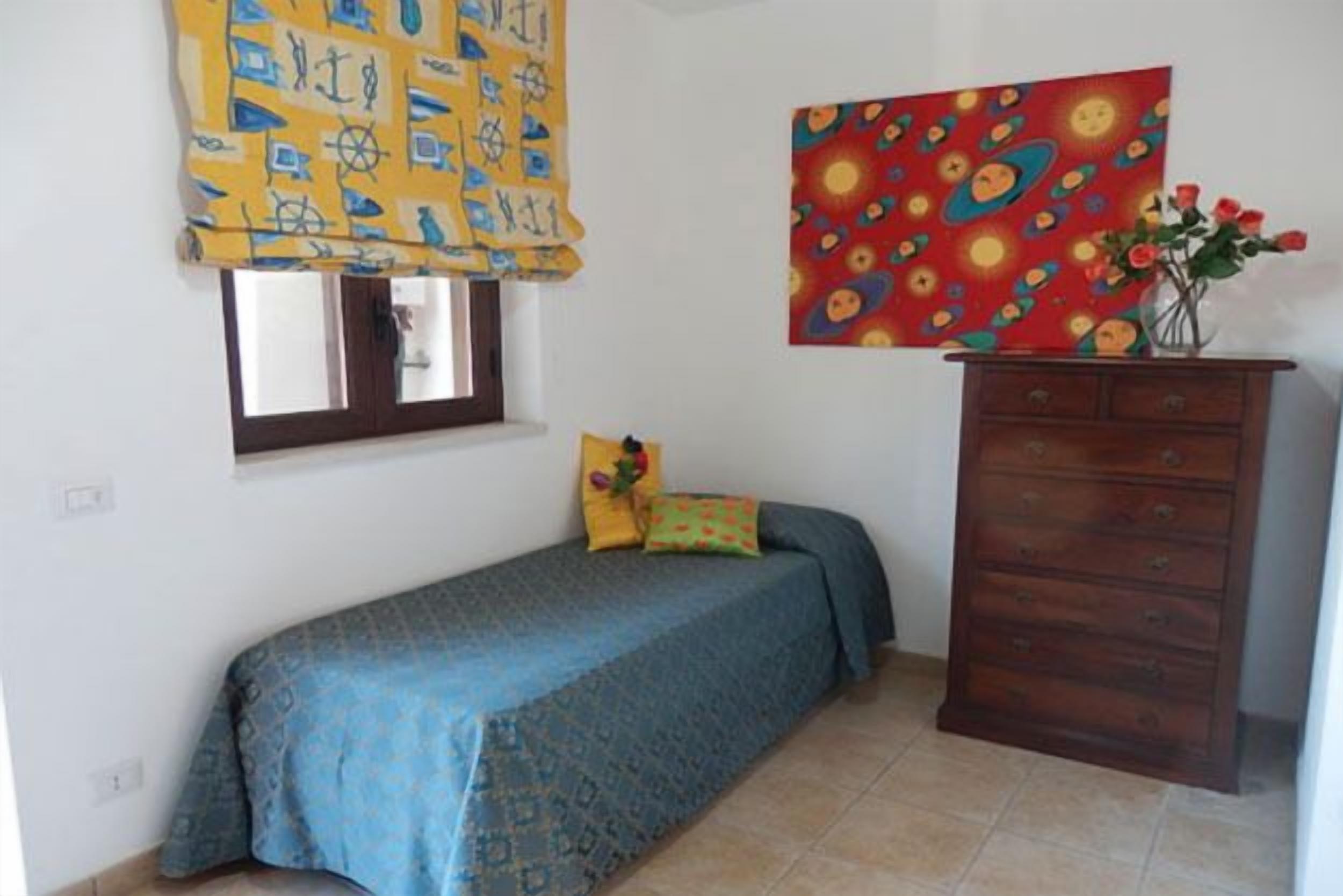 A small bedroom with a single bed, a wooden dresser, a window with curtains, and a colorful painting on the wall.