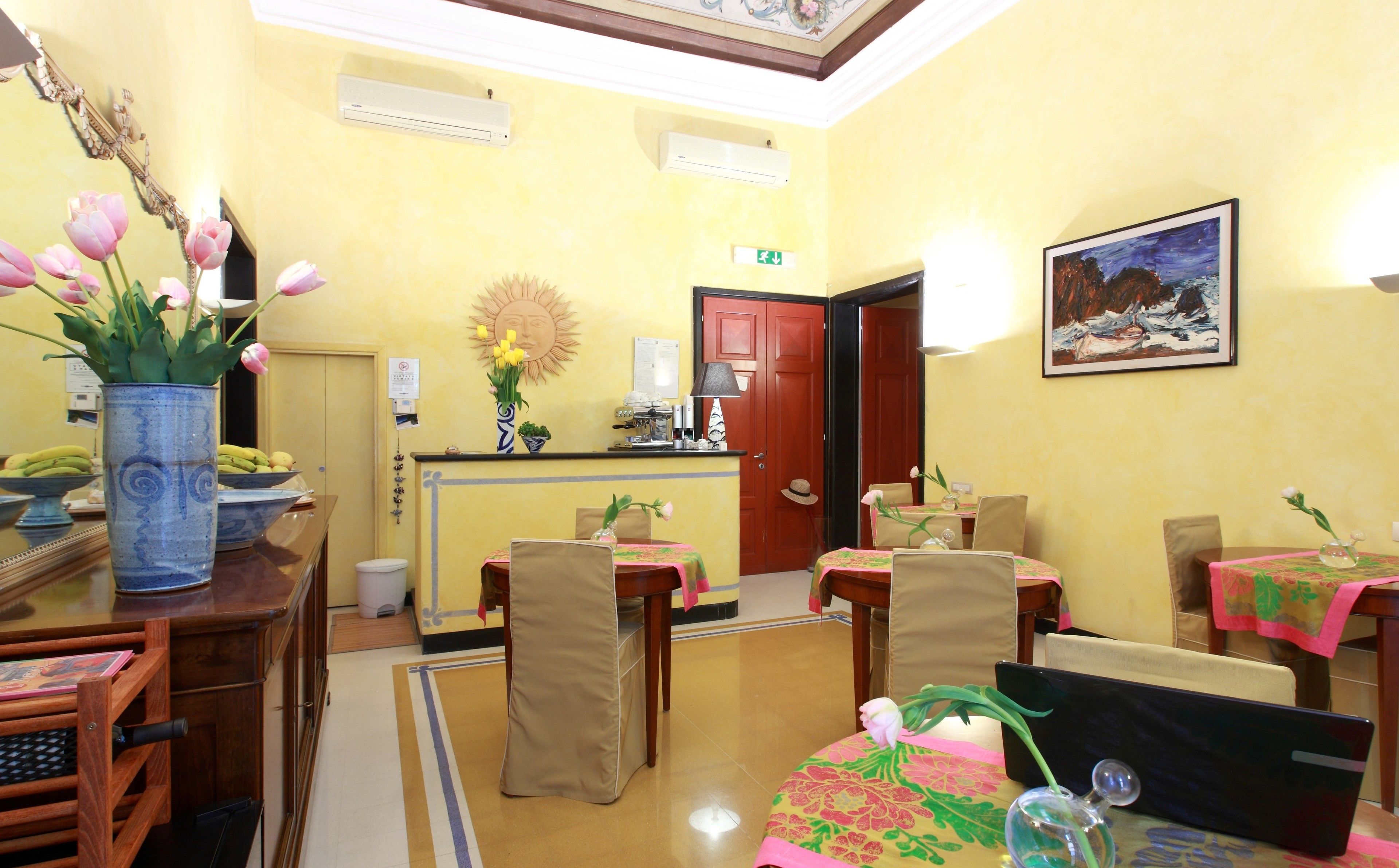 A hotel lobby with a reception desk, a coffee machine, a dining area with tables and chairs, and a painting on the wall.