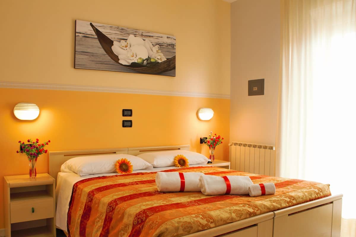Un cuarto de hotel con cama, dos mesitas de noche, una lámpara, un florero con flores, un cuadro en la pared y una ventana con cortinas.
